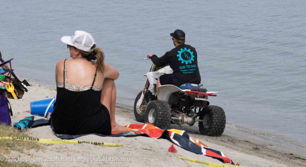 A person sitting on the sand watches another person ride an ATV into shallow water.