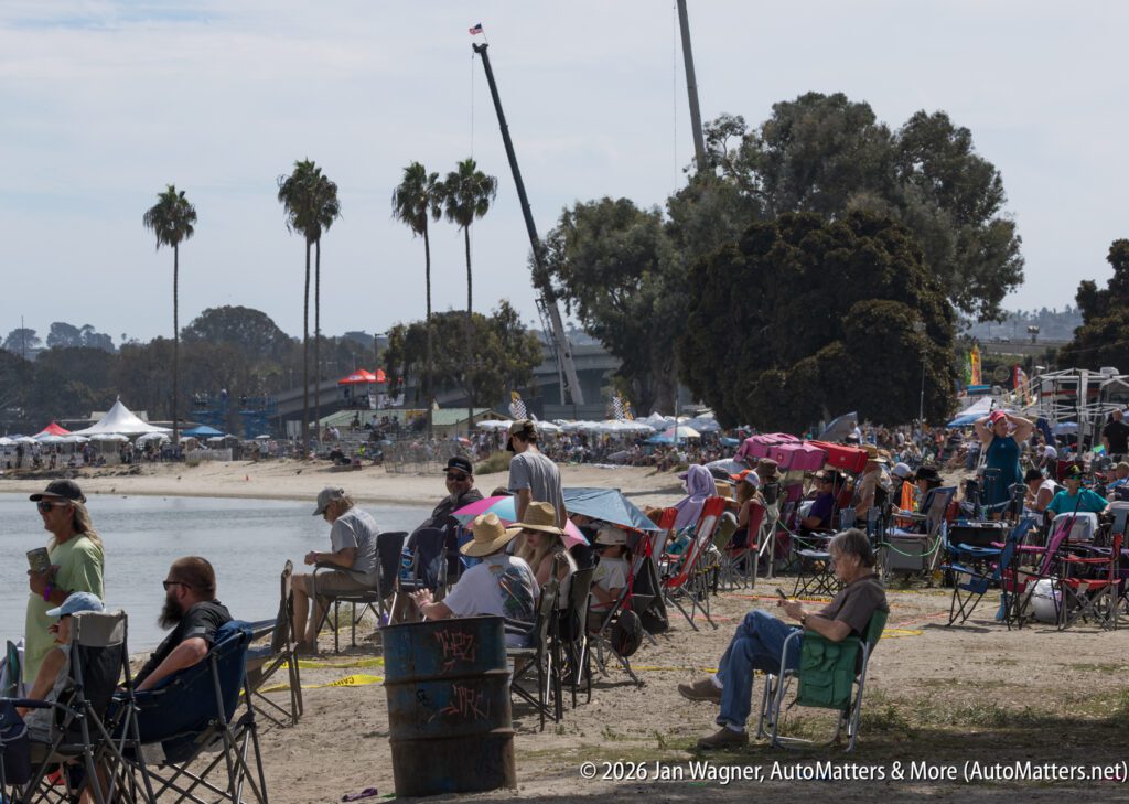 Spectators in folding chairs sit along a sandy shore watching a public event.