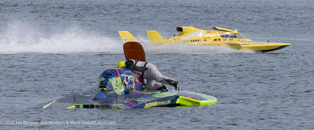 A driver crawls onto their capsized, multicolored hydroplane in the water as a yellow race boat speeds by in the background.