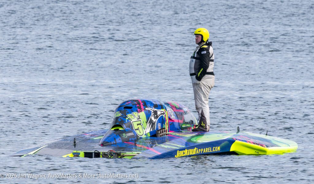 A person in a helmet and jumpsuit stands on a colorful, partially submerged racing boat in the water.