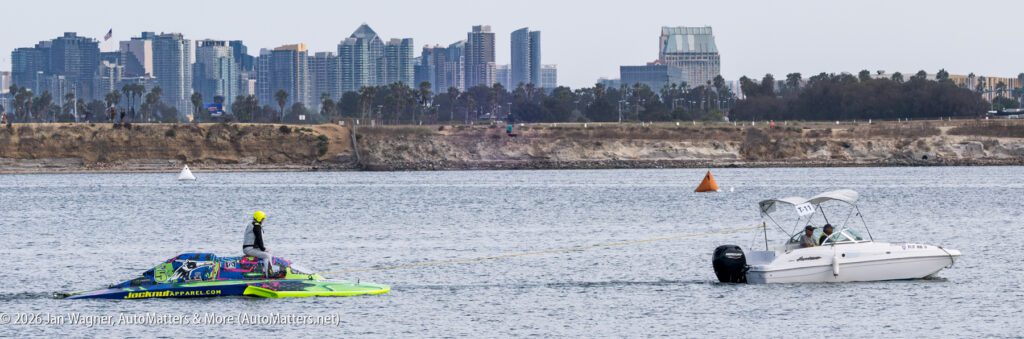 A white motorboat tows a bright green and blue race boat with a yellow-helmeted driver on a bay with a city skyline in the background.