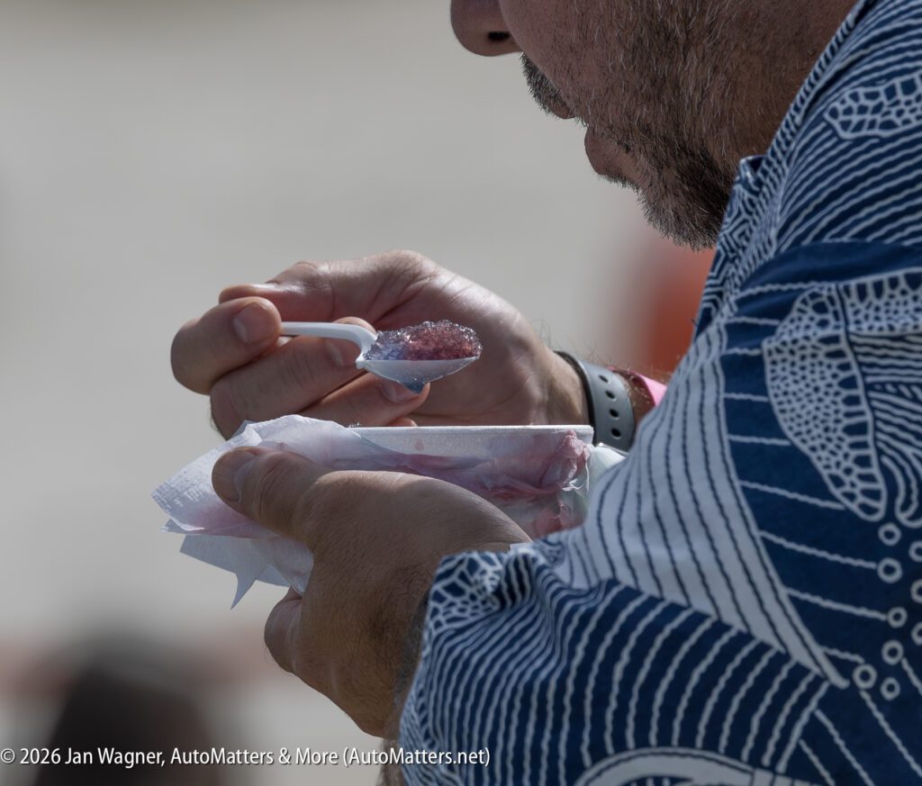A person wearing a blue patterned shirt eating purple shaved ice with a plastic spoon.