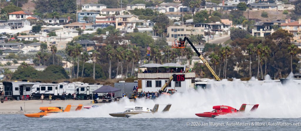 Three hydroplane race boats speeding across a body of water, creating large spray plumes in front of a shore with motorhomes and houses.