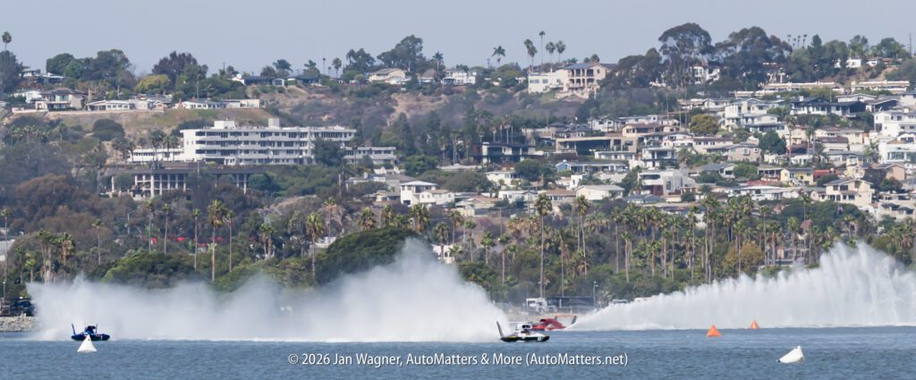 Two hydroplane race boats speeding across a bay, creating large plumes of water spray, with a residential hillside in the background.