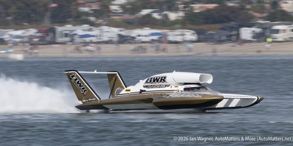 A gold and white BWR Racing hydroplane boat speeding across the water, creating a large white wake.