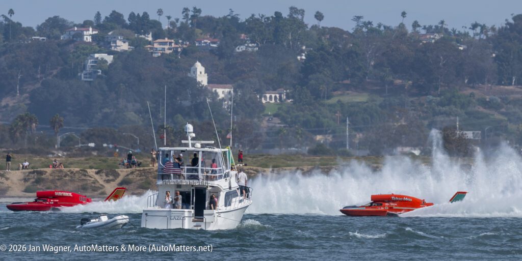 Two racing hydroplanes speed through the water alongside a large motor yacht on a bay.