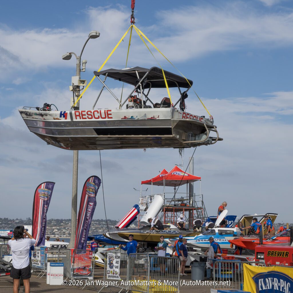 A crane lifts a metal rescue boat labeled “RESCUE” above a busy race boat pit area.