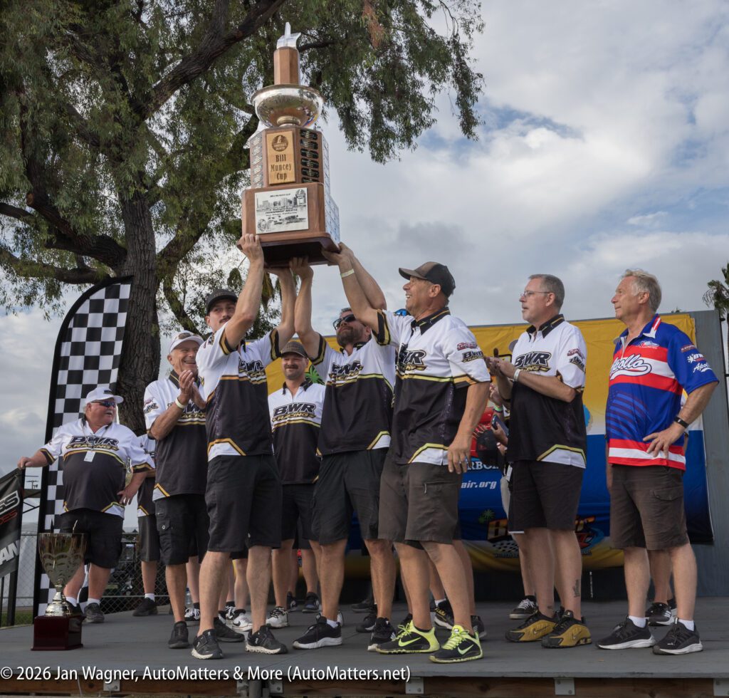 A racing team lifts a large trophy, the Bill Munces Cup, while standing on a podium.