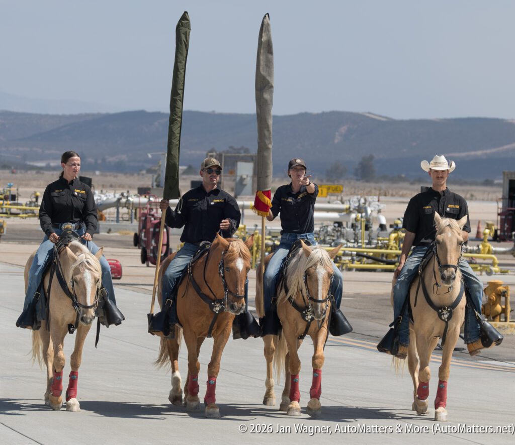 Four mounted riders in black shirts and jeans carry vertical banners while riding horses on an airfield.