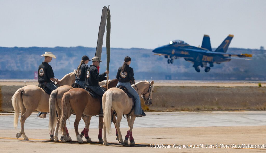 Three riders on horseback carry flags while a Blue Angels F/A-18 Hornet jet flies in the background.