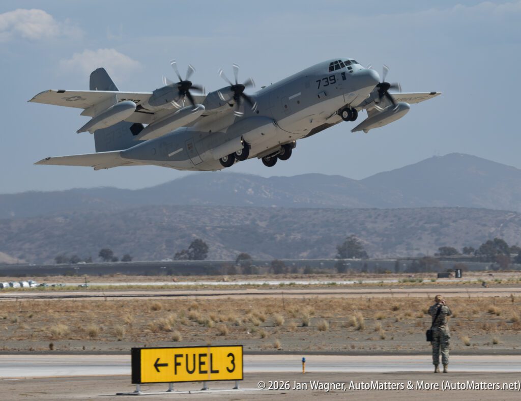A gray U.S. Marine Corps KC-130J Hercules aircraft taking off, with a person in camouflage uniform standing nearby on the airfield.