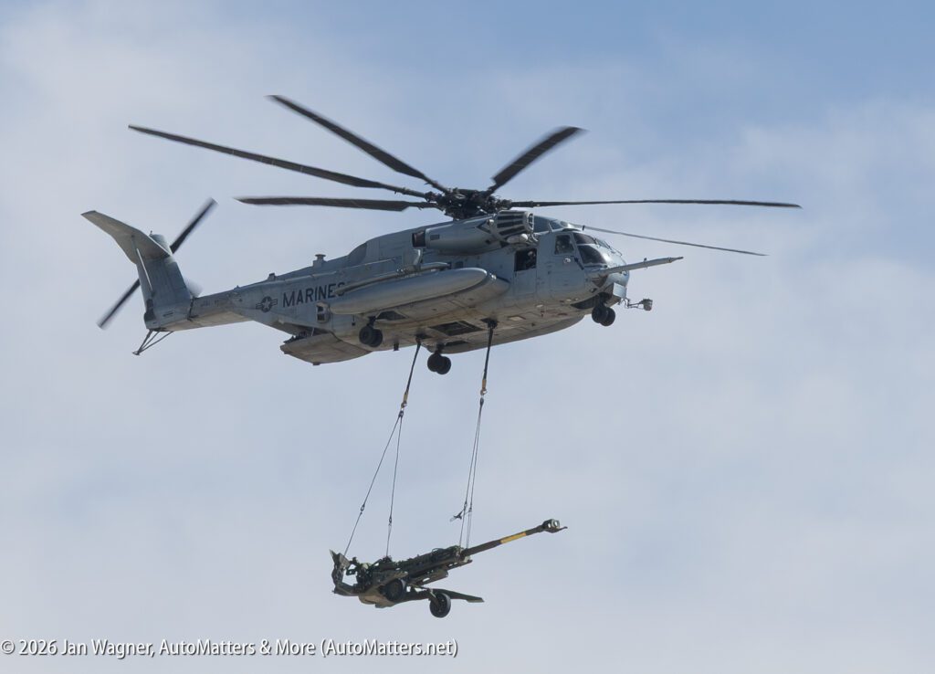 U.S. Marine Corps CH-53E Super Stallion helicopter carrying an artillery piece slung beneath it in flight.