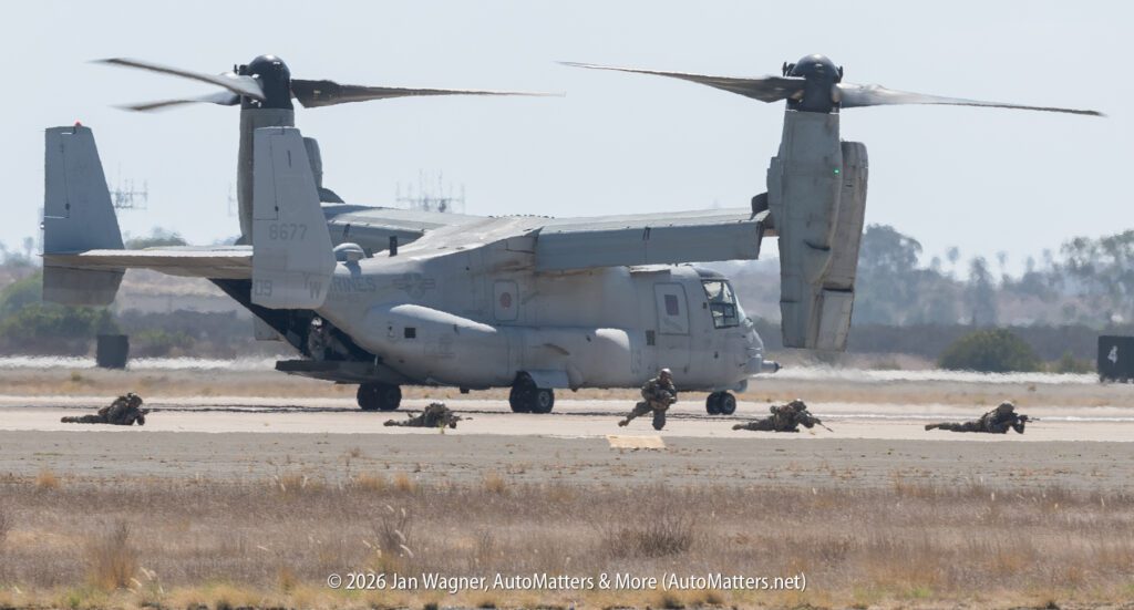 U.S. Marines in combat gear spread out on an airfield in front of a parked V-22 Osprey tiltrotor aircraft.