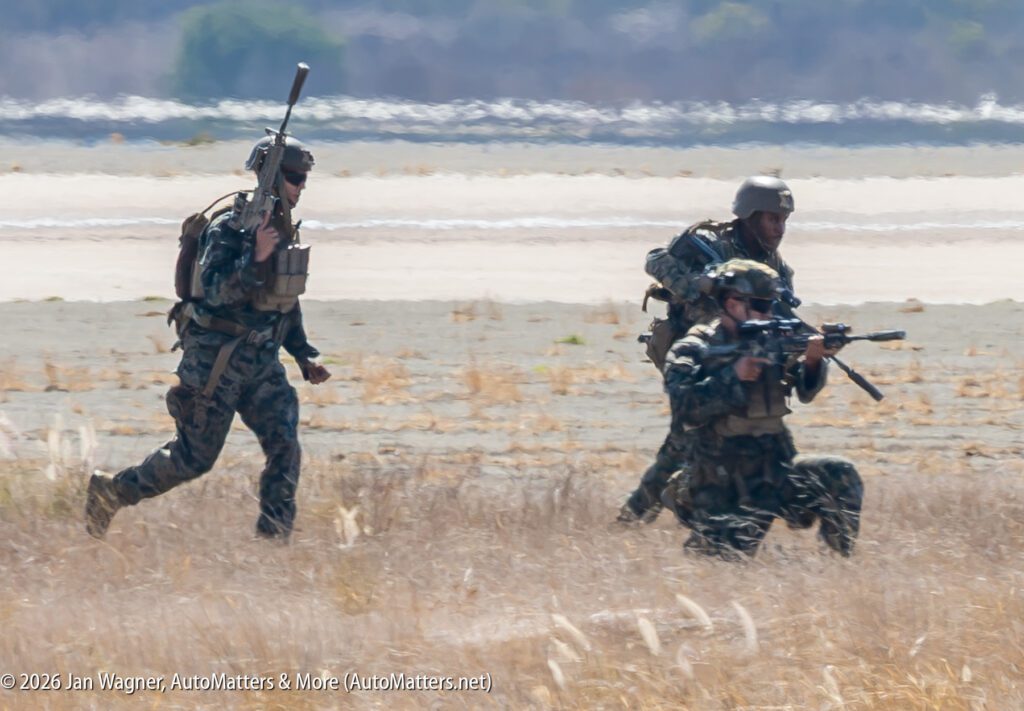 Three soldiers in camouflage uniforms and helmets move through a dry field, with two holding rifles.