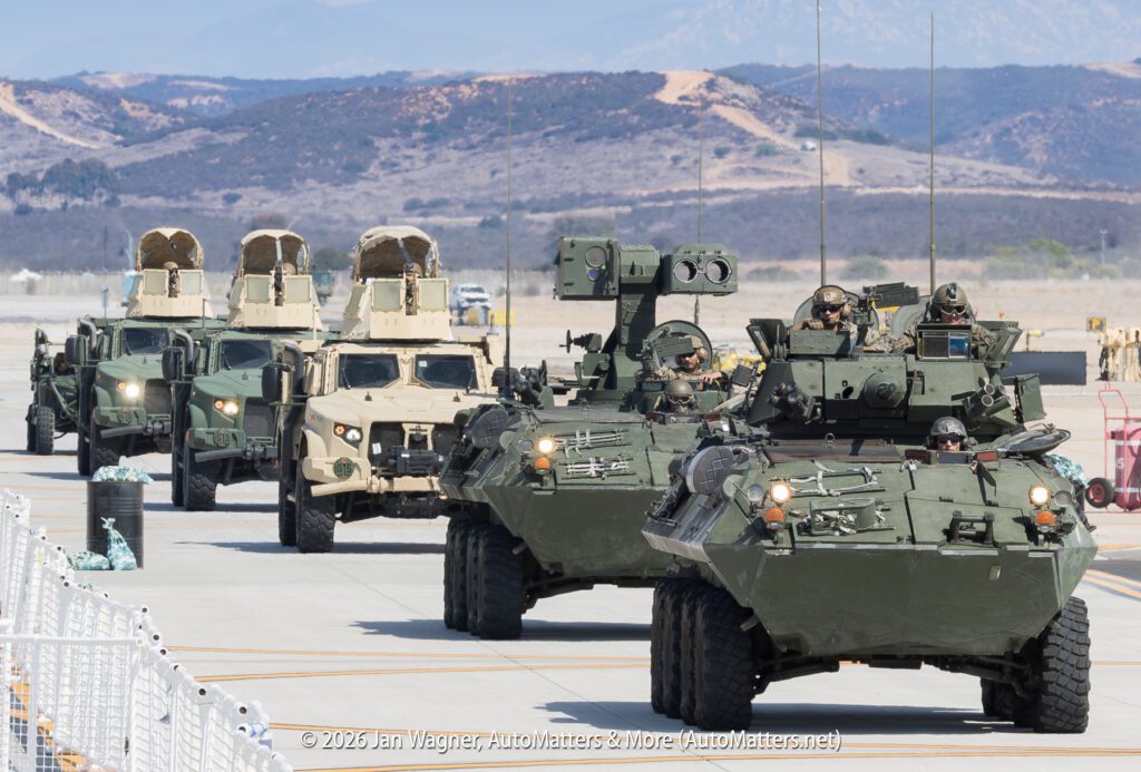 A line of military vehicles, including armored personnel carriers and light tactical vehicles, driving on a paved lot.
