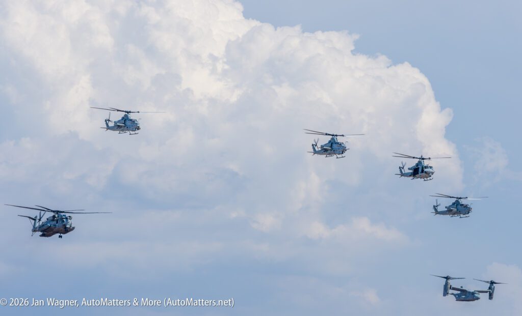 Five military helicopters and an Osprey aircraft flying in formation against a cloudy sky.