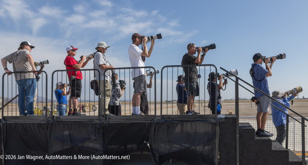 A group of photographers stands on an elevated platform, using long-lens cameras to photograph an airfield.