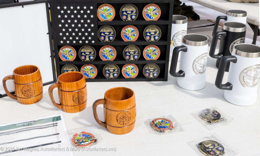 A wooden display case holds rows of military challenge coins, flanked by three wooden beer steins and two white travel mugs on a table with price lists and individual coins.