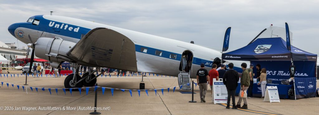 A vintage silver and blue United DC-3 airplane parked on an airfield with a display tent and people gathered nearby.