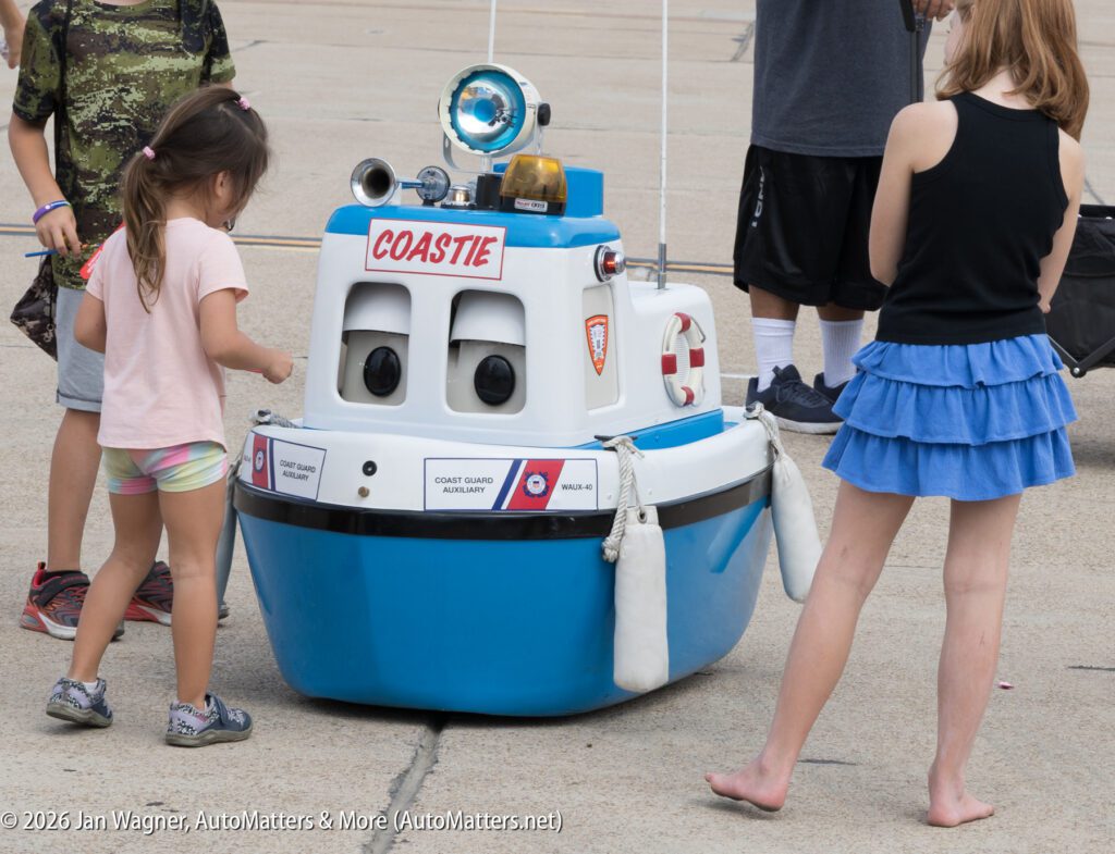 Two children stand near a Coast Guard Auxiliary “Coastie” mascot boat on wheels.