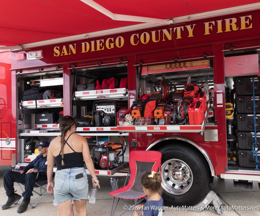 A woman and child stand before a San Diego County Fire truck with its equipment compartments open, revealing tools, cases, and a generator.