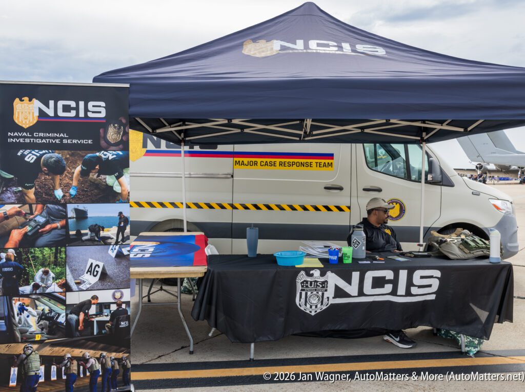 A man in an NCIS polo shirt sits at a branded information booth in front of an NCIS Major Case Response Team van.