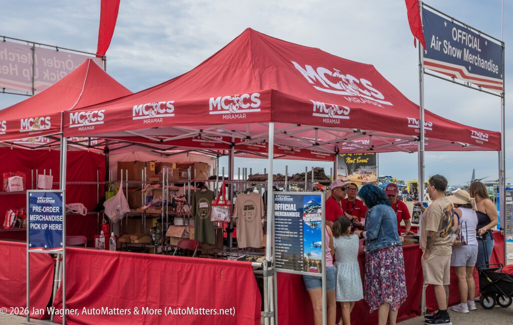A red tent selling official air show merchandise, with a price list sign and people browsing the items.