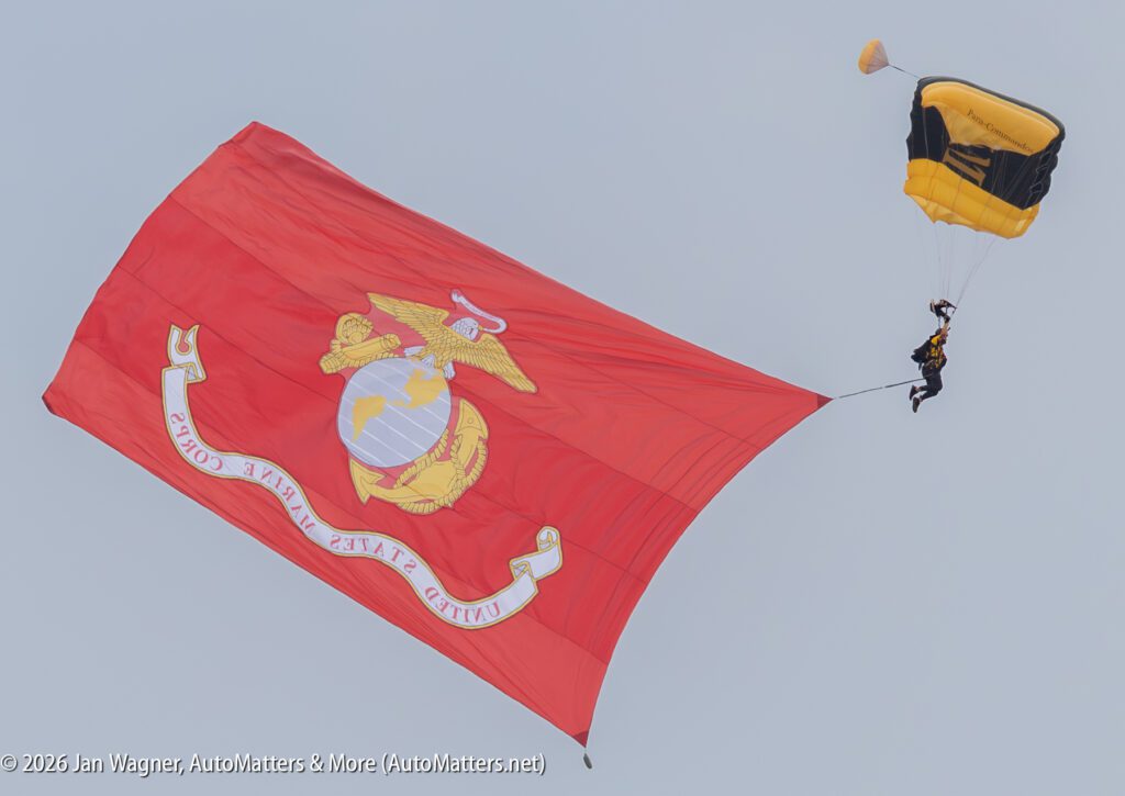 A skydiver with a black and yellow parachute descends while trailing a large United States Marine Corps flag.