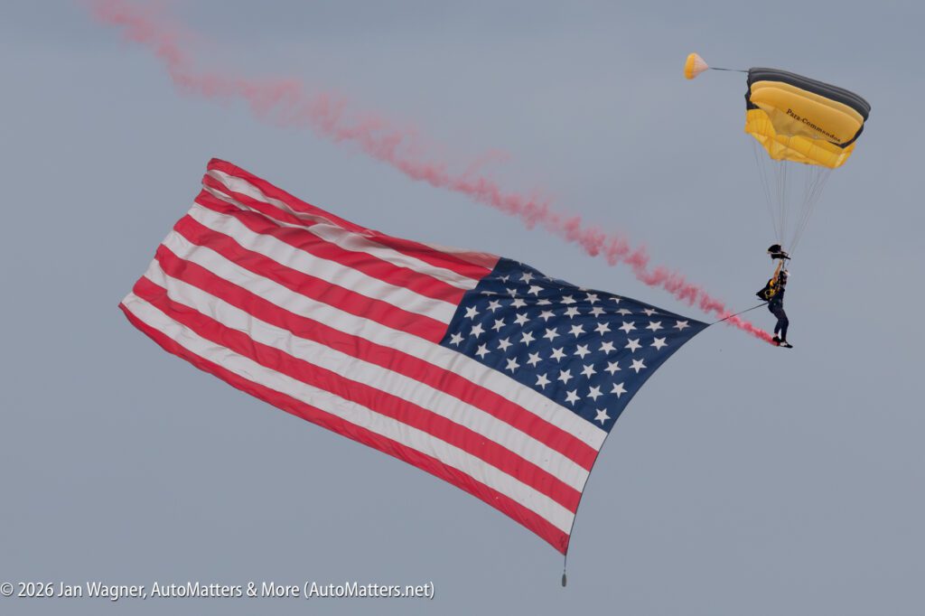 A skydiver with a yellow parachute trailing red smoke carries a large American flag.