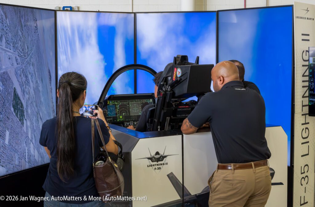 People observe a pilot seated in an F-35 flight simulator with three large screens displaying aerial views.