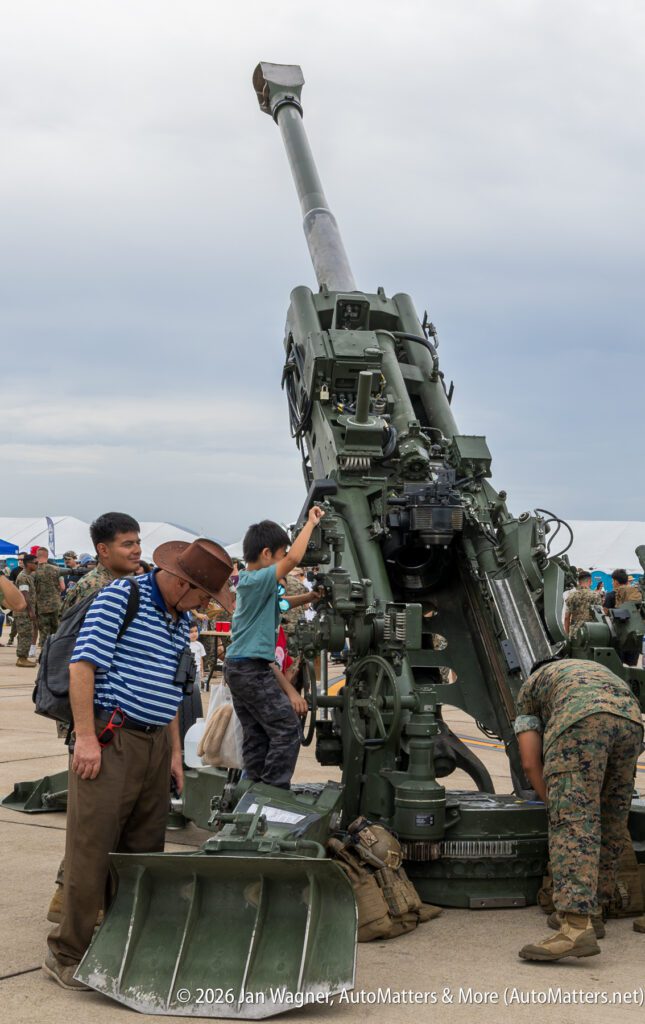 A child and two men examine a large military artillery piece on a paved lot.