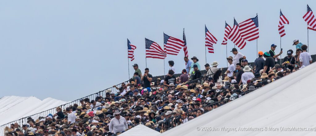 A large crowd sits in stadium bleachers under a row of American flags.
