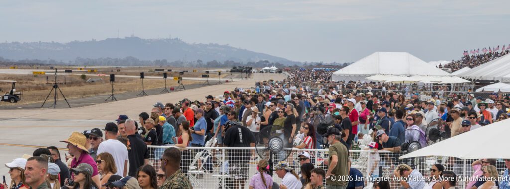 A large crowd of people standing behind a fence, looking toward an airfield with mountains in the background.