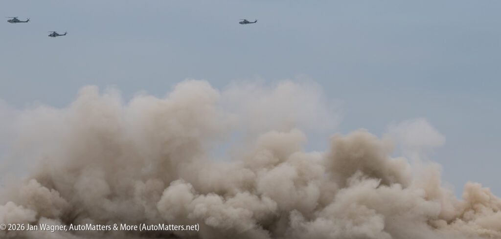 Three military helicopters fly above a large cloud of dust.