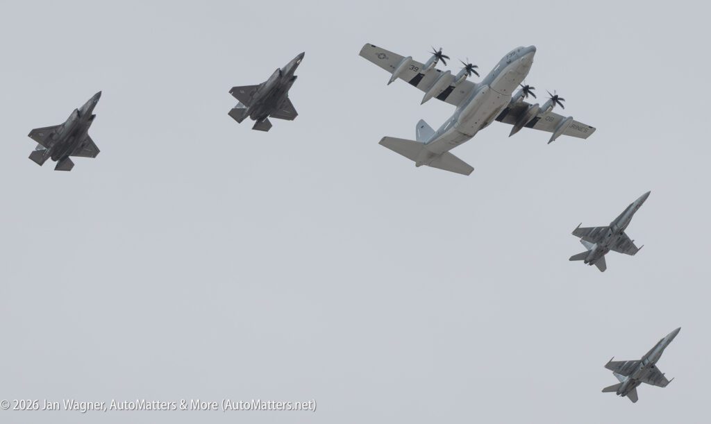 A C-130 Hercules transport aircraft flying in formation with four fighter jets against a cloudy sky.