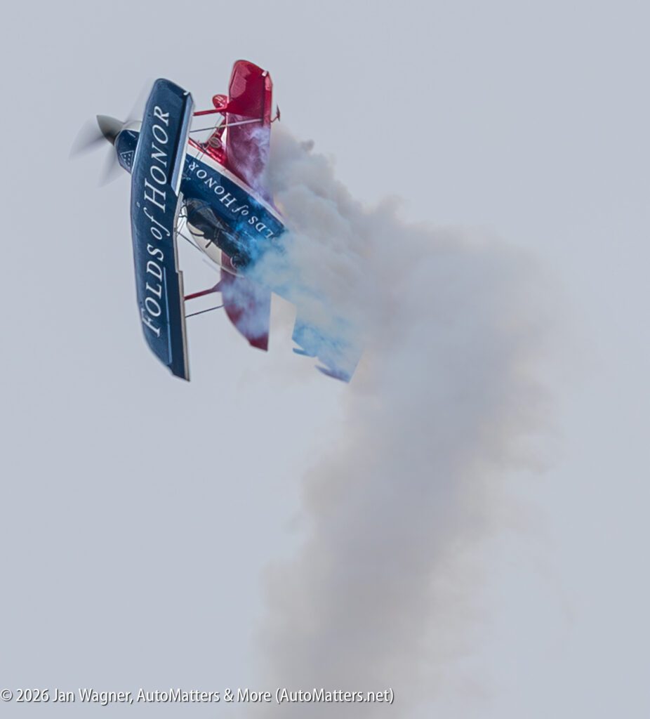 A red and blue stunt biplane labeled “Folds of Honor” performing a maneuver with a smoke trail.