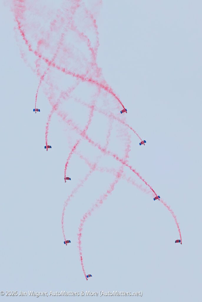 Seven skydivers with blue and red parachutes descend, trailing long ribbons of red smoke in the sky.