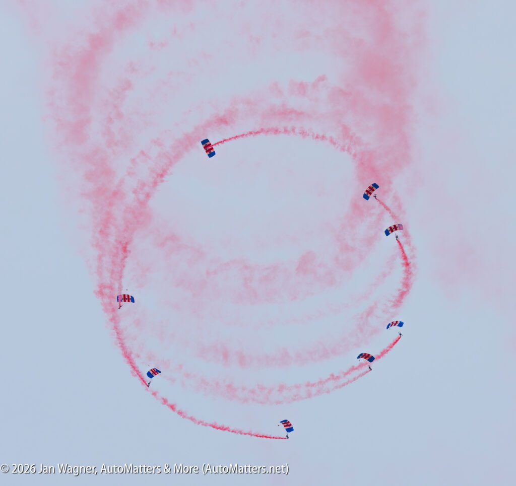 Seven skydivers descending in a circle, each trailing red smoke against a cloudy sky.