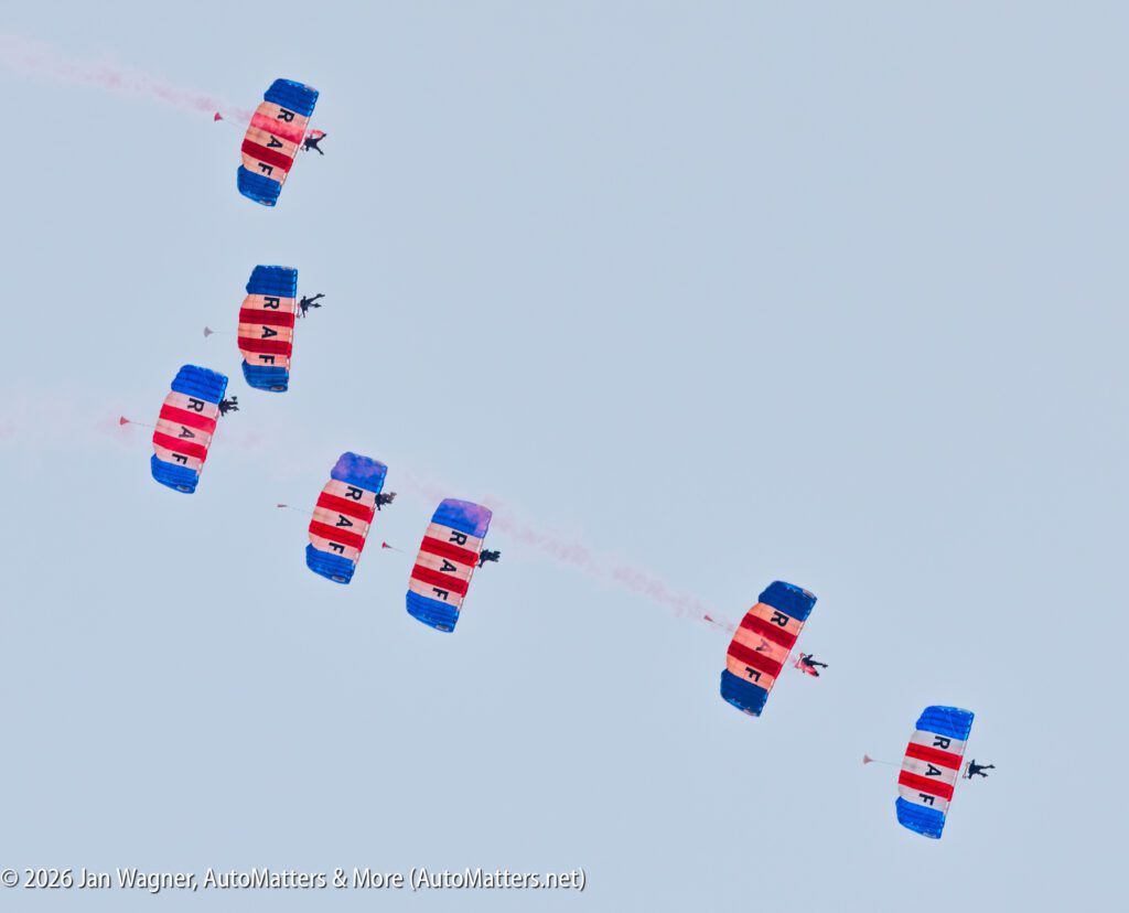 Six skydivers in formation, each using a parachute with red and white stripes and blue “RAF” lettering, descending against a clear sky.