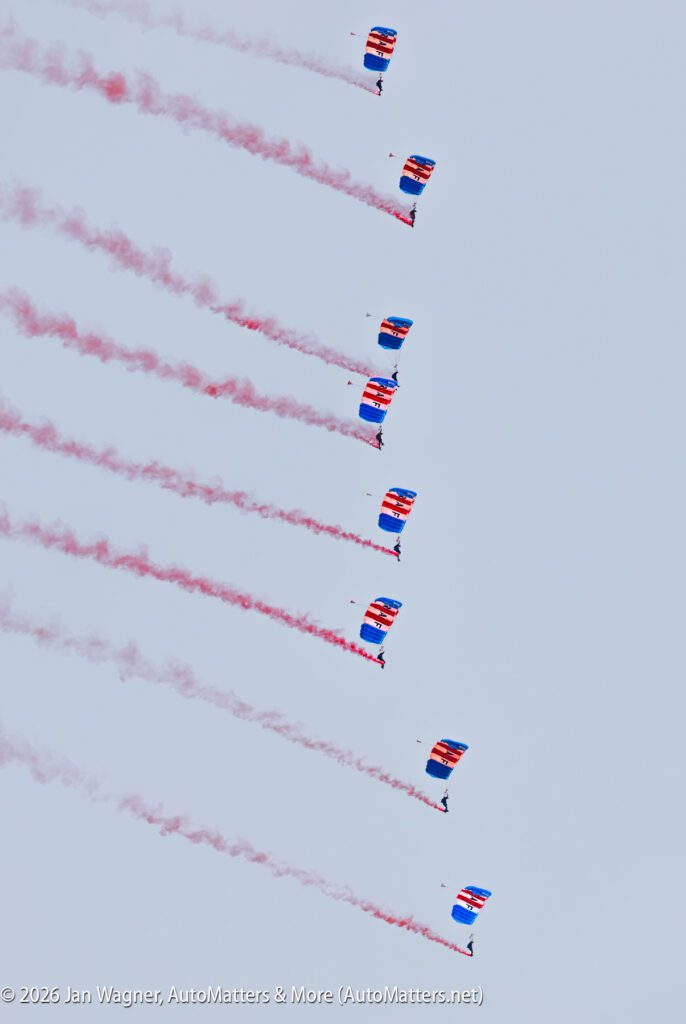 Seven skydivers in a diagonal formation, each using a red, white, and blue parachute and trailing red smoke against a clear sky.