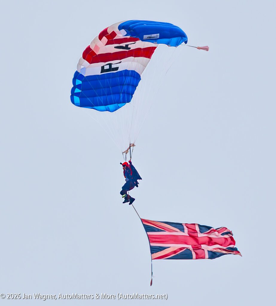 A skydiver descends with a red, white, and blue parachute, trailing a large Union Jack flag below them.
