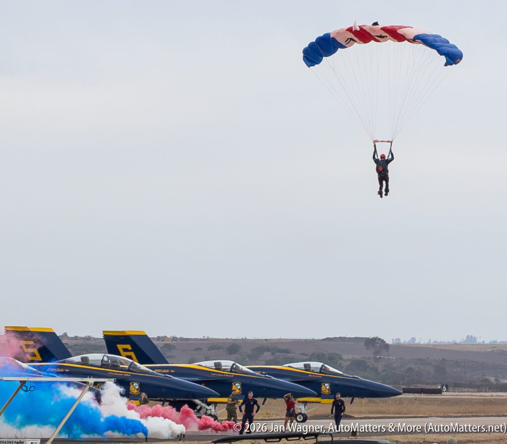 A parachutist descends above three parked Blue Angels fighter jets emitting red and blue smoke.