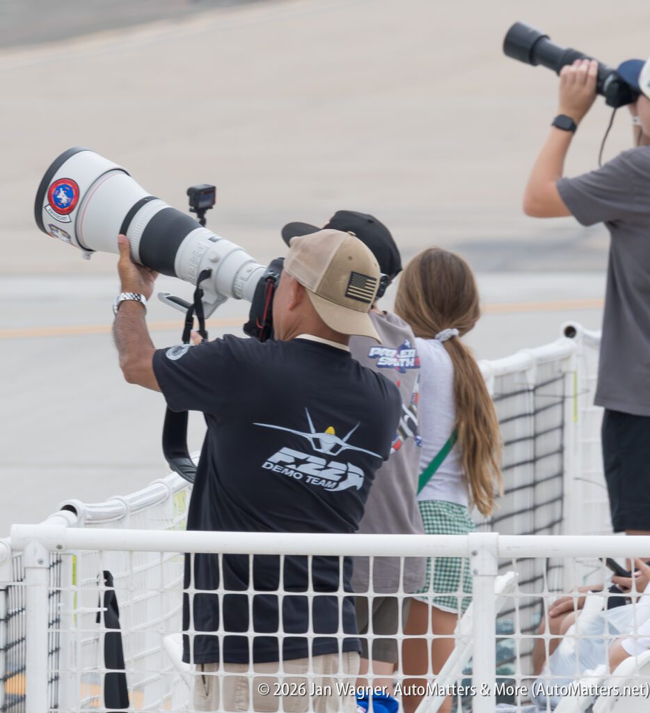 A photographer in an F-22 Demo Team shirt aims a large telephoto lens at an air show, standing beside two other people with cameras behind a safety fence.