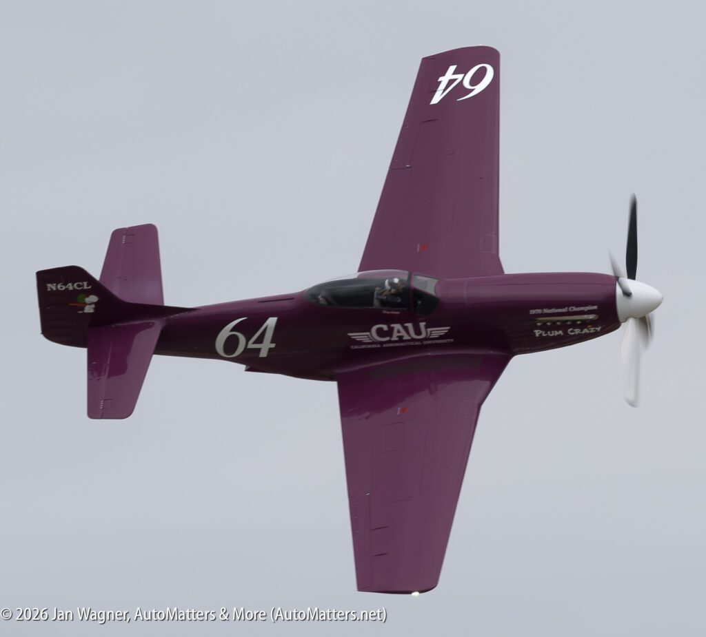 A purple P-51 Mustang aircraft, named “Plum Crazy,” in flight against a gray sky.