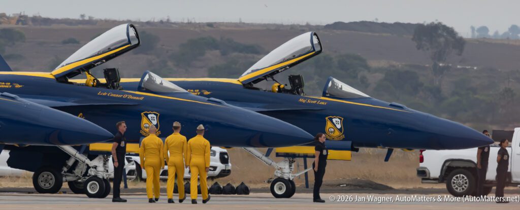 Three Blue Angels F/A-18 Hornet jets parked on a runway with ground crew in yellow jumpsuits and black uniforms standing nearby.