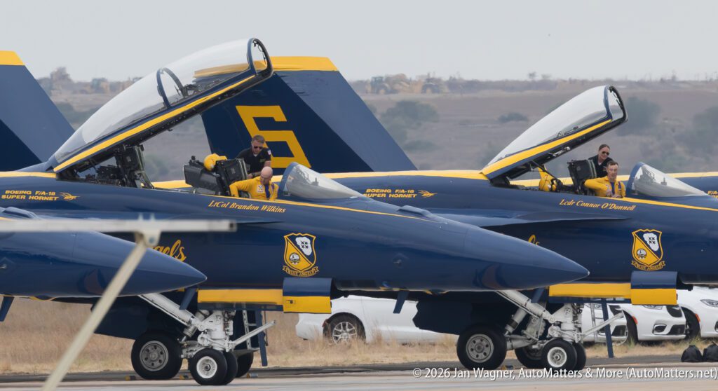Two Blue Angels F/A-18 Super Hornet fighter jets parked on a flight line with their canopies open and pilots seated in the cockpits.