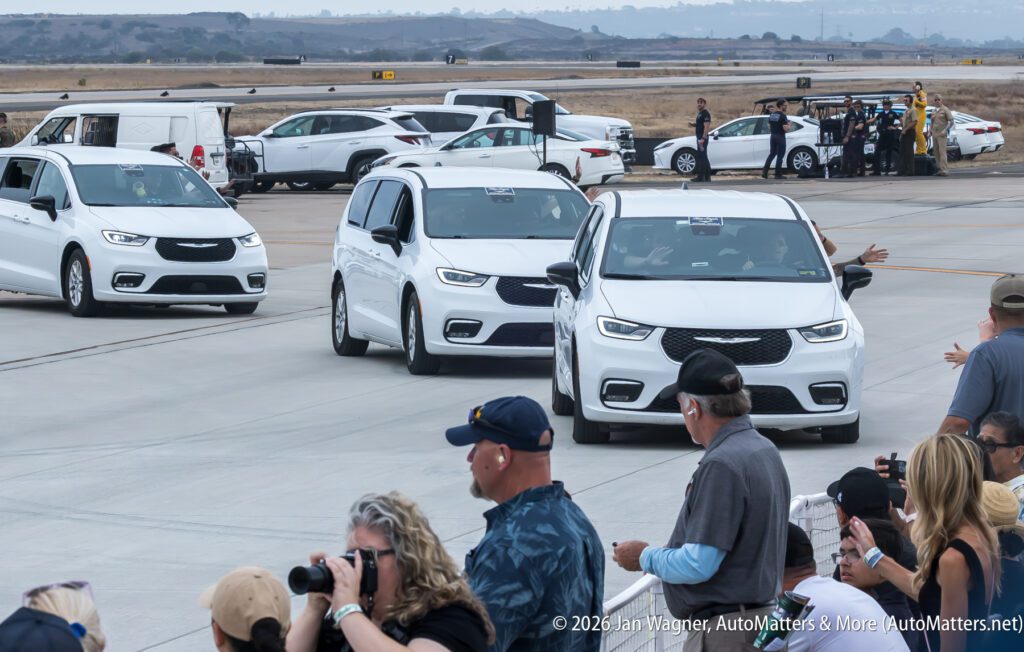 A line of white Chrysler Pacifica minivans driving past a group of onlookers at an airfield.