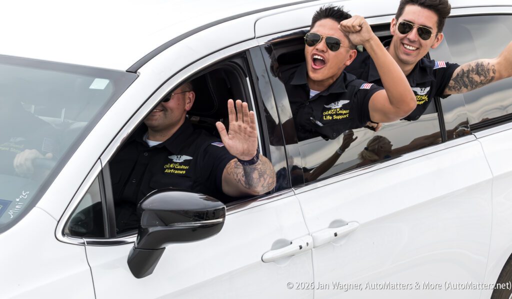 Three people in matching black shirts waving from the windows of a white SUV.