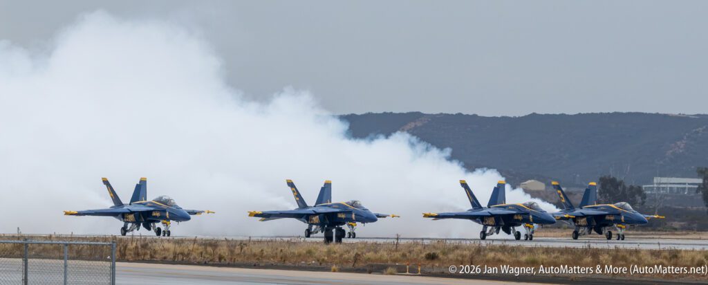 Four U.S. Navy Blue Angels F/A-18 Hornet jets taxi on a runway with smoke trails behind them.