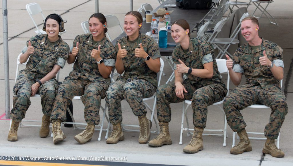 Five U.S. Marines in camouflage uniforms sit in a row of white folding chairs, smiling and giving thumbs-up gestures.